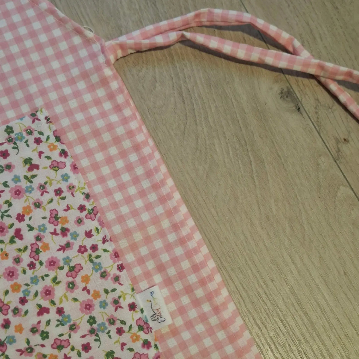 Detalle de una estola de cuadros rosas con bolsillo floral y cinta de ajuste sobre una mesa de madera.