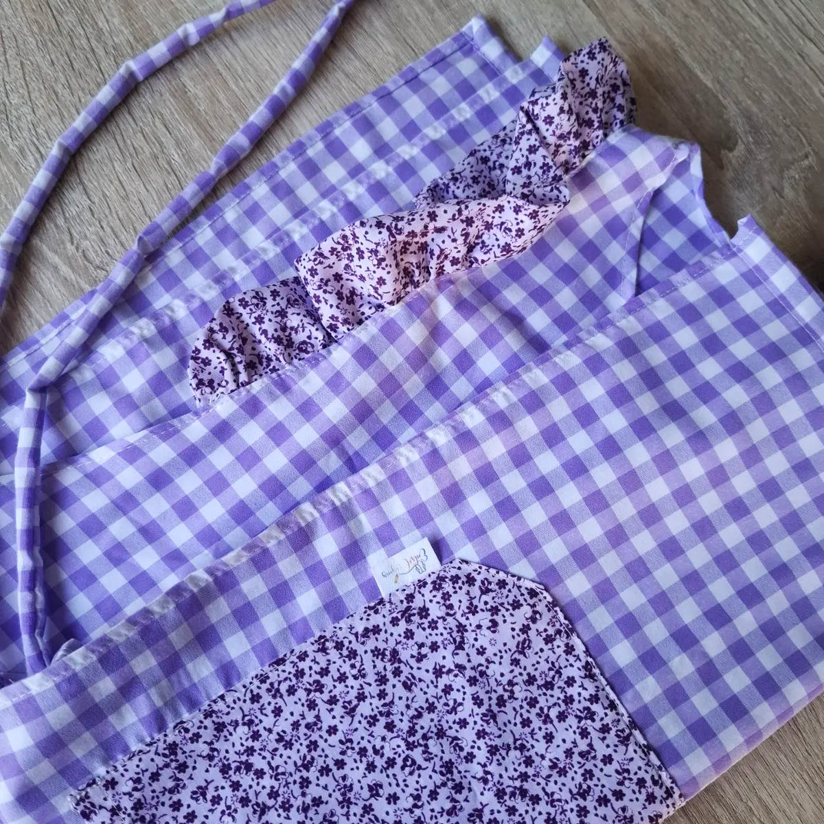 Detalle de una estola de cuadros lavanda con volante floral, bolsillo y cintas de ajuste.